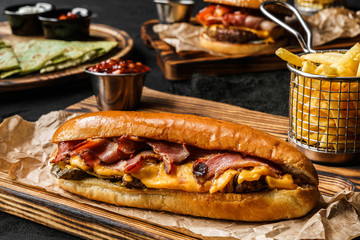 Canvas Print - Tasty burger sandwich with meat cutlet, slices of cheese, ham and pickles on wooden board with french fries potatoes on black background. Fast food lunch, close up