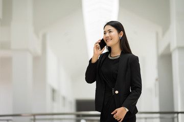 Portrait of a young Asian business woman talking over cellphone and holding cup of coffee in business building. Photo of beautiful girl in casual suite with phone and cup of coffee.