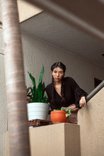 From Below Of Stylish Ethnic Lady With Vitiligo Skin Condition In Black Jacket Sitting Near Potted Plants In Apartment While Looking At Camera