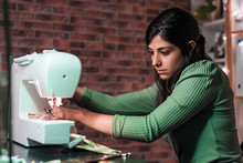 Side View Of Craftswoman Using Modern Sewing Machine While Creating Soft Fabric Samples With Creative Green Pattern Near Lamp In Loft Style Workshop