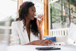© Fernando Valencia/ADDICTIVE STOCK - Low angle of black confident woman drinking water from glass while using netbook during work in cozy workplace in cafe