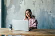© Ilona Titova/ADDICTIVE STOCK - Young female remote employee working on netbook while sitting at wooden table with disposable glass of coffee near old cement wall in flat