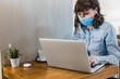 © Israel De Lago/ADDICTIVE STOCK - Focused female freelancer in denim shirt and medical mask using earbuds and working remotely in cafe using laptop during coronavirus epidemic