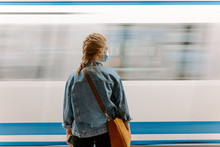 Back View Of Millennial Female Passenger In Protective Mask And Casual Denim Jacket With Handbag Standing On Underground Platform In Front Of Moving Train