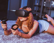 © Jota Gil/ADDICTIVE STOCK - Seductive barefoot anonymous female in casual wear lying with open mouth on soft plaid on bed while playing game with controllers and in goggles of virtual reality
