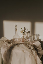 High Angle Of White Delicate Flowers In Small Vase Placed On Table Covered With Tulle And Arranged With Vintage Styled Glasses And Decanter