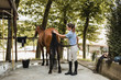 © Raquel Arocena/ADDICTIVE STOCK - Side view of busy female rider standing near barn and grooming obedient horse with brush