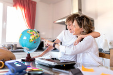 Positive Boy Cuddling Gentle Happy Woman Pointing With Finger At Colorful Globe While Sitting At Table In Kitchen And Explaining Geography Lesson To Son At Home