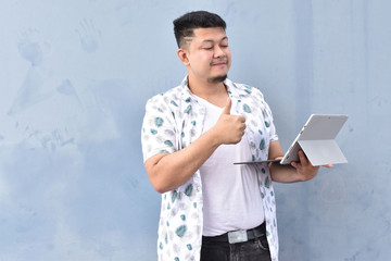 Indoor portrait of young man in t shirt standing against textured wall with copy space for ads, holding laptop and looking at camera with happy smile