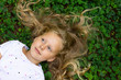 © uladzimirzuyeu - Child girl with blond hair on green grass background. A child with a smile lies on the grass in the park and smiles.