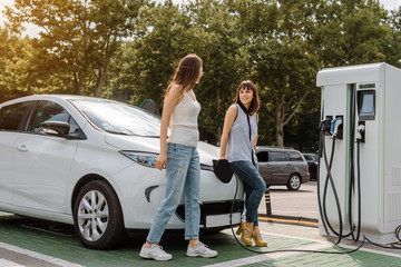 Two happy young beautiful women are talking to each other until their electric car is charging at the charging station situated in the car park.