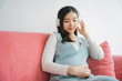 © THESHOTS.CO - Young beautiful pregnant asian woman listen to song with headphone on a couch.