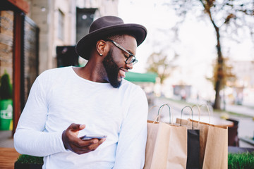 Happy afro american guy in trendy hat getting package with purchases from online shop sending good feedback on mobile, cheerful dark skinned man satisfied with buying checking mock up paper bags