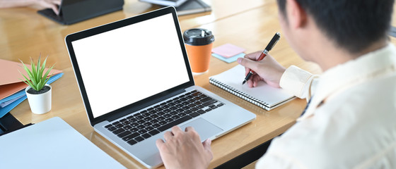 Wall Mural - A man is using a white blank screen laptop while taking notes and sitting at the wooden table.