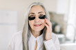 © sofiko14 - Close up beauty portrait of smiling pretty senior gray haired woman, applying under eye mask, black patches on her face with hands, posing to camera on the background of home interior