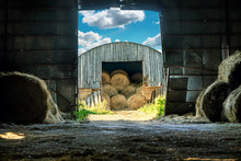 View Of The Hay Bales Being Stored In An Old Wooden Barn From The Another Hay Barn In Late Summer Or Early Autumn Sunny Evening. Rural Scene
