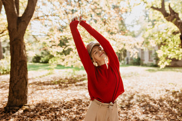 Wall Mural - Beautiful stylish girl smiling in the park under sun. Charming blonde in red sweater feeling happy outdoor.