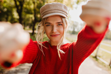 Wall Mural - Lovely blonde girl in nice trendy hat and red pullover making selfie in the autumn park.