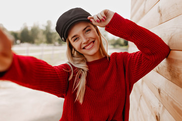 Wall Mural - Lovely blonde in red sweater and black hat cheerfully smiling at her phone. Beautiful woman making selfie near wooden house in park.