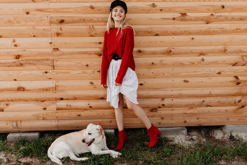 Wall Mural - Smiling girl enjoys nice autumn day posing on the wooden background. Happy blonde standing on the green grass near her white dog.