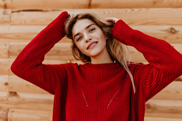 Young blonde woman in red sweater standing near wooden house. Beautiful blonde playing with her hair outdoor.