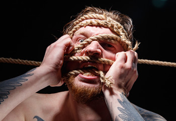 Image of binded tattooed man trying tear the rope on face