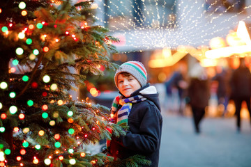 Wall Mural - Little cute kid boy having fun on traditional Christmas market during strong snowfall. Happy child enjoying traditional family market in Germany. Schoolboy standing by illuminated xmas tree.