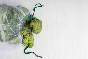 Wall Mural - Top view of transparent net bag with green raw broccoli on white wooden background showing sustainable consumption and environmental protection. Image with copy space, horizontal orientation