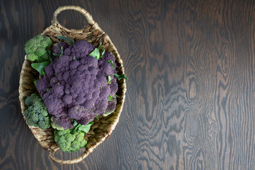 Wall Mural - Top view of lilac cauliflower or purple broccoli in fruit basket on dark brown wooden background at kitchen ready for cooking. Image with copy space, horizontal orientation