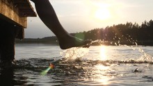 Persons Feet Splashing Water At The Sunset. Enjoying Holidays By Lake