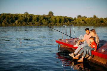 Happy young family fishing on pier on river or lake in summertime. Photography for ad or blog about family and travel