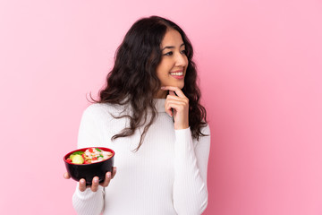 Wall Mural - Mixed race woman holding a bowl full of noodles over isolated pink background thinking an idea and looking side