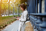Fototapeta Konie - Young beautiful woman is booking on-line via portable touch pad, while is standing on the street in autumn day. Female dressed in spring clothes is reading information in internet via digital tablet