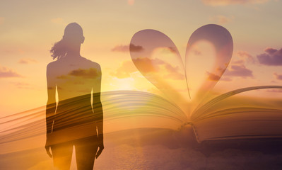 Female silhouette facing the sunset next to bible book with page shaped into heart. Love, religion, and spiritual concept. 