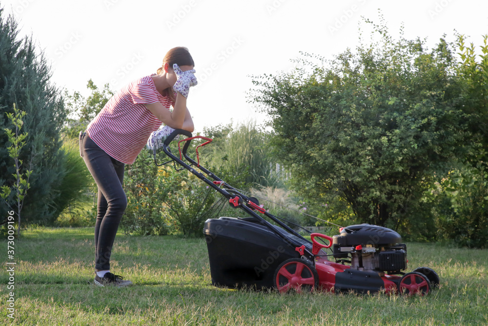 Tired woman making a short break while cutting grass with a lawn mower. Outdoor household chores concept. - obrazy, fototapety, plakaty 