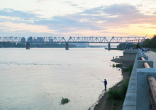 Silhouette Of A Lonely Fisherman With A Fishing Rod On The River Bank Near The Embankment In The Evening Against The Background Of The Railway Bridge.