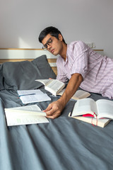 Wall Mural - Vertical shot of a young male with glasses studying on his bed with books and textbooks on it