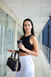 © Natalia - Young brunette woman, wearing white pants and black top, holding black purse, cell phone and paper coffee cup, standing in light office building. Businesswoman on lunch break.