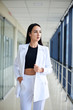 © Natalia - Young brunette woman, wearing white pants and jacket, standing in light passageway with huge windows, posing for social media. Businesswoman on a break. Female portrait photography.