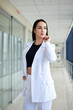 © Natalia - Young brunette woman, wearing white pants and jacket, standing in light passageway with huge windows, posing for social media. Businesswoman on a break. Female portrait photography.