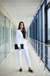 © Natalia - Young brunette woman, wearing white pants and jacket, standing in light passageway with huge windows, holding black clutch purse, posing for social media. Businesswoman on a break. Female portrait.