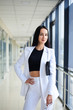 © Natalia - Young brunette woman, wearing white pants and jacket, standing in light passageway with huge windows, holding black clutch purse, posing for social media. Businesswoman on a break. Female portrait.
