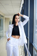 © Natalia - Young brunette woman, wearing white pants and jacket, standing in light passageway, posing for social media, leaning on window. Businesswoman on a break. Female portrait photography.
