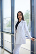 © Natalia - Young brunette woman, wearing white pants and jacket, standing in light passageway with huge windows, posing for social media, leaning on railing. Businesswoman on a break. Female portrait photography