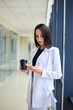 © Natalia - Young brunette woman, wearing white business suit, holding cell phone and black paper coffee cup, standing in light office building near window. Businesswoman on lunch break. Corporate culture concept