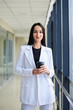 © Natalia - Young brunette woman, wearing white business suit, holding black paper coffee cup, standing in light office building near window. Businesswoman on lunch break. Corporate culture concept.