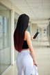 © Natalia - Young brunette woman, wearing white pants and black top, holding jacket in hand, looking at cell phone in light passageway of office building. Businesswoman on lunch break. Corporate culture concept.