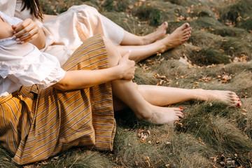 Women sitting on dry grass with fallen leaves on a warm autumn day, close-up on barefoot legs.