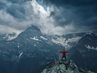 Alone hiker man in red jacket against the gloomy mountain top landscape with thunder cloudy sky, rocky ranges and peaks with glaciers and snow fields. Domestic travel and trekking. Local tourism.