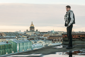 Wall Mural - A bearded guy in a leather vest and jeans stands on the edge of the roof with a view of St. Petersburg at sunset